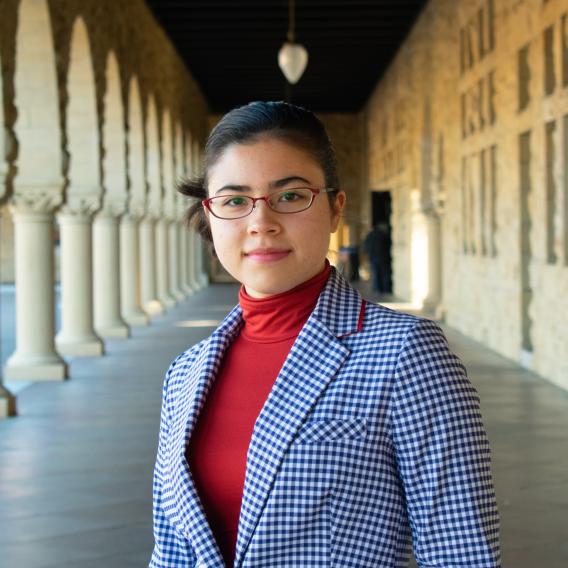 Picture of Mei Li. In the picture, she is looking at the camera with a soft smile. Mei Li is wearing red glasses and she is wearing a blue and white checkered blazer with a bright red turtle neck underneath. 