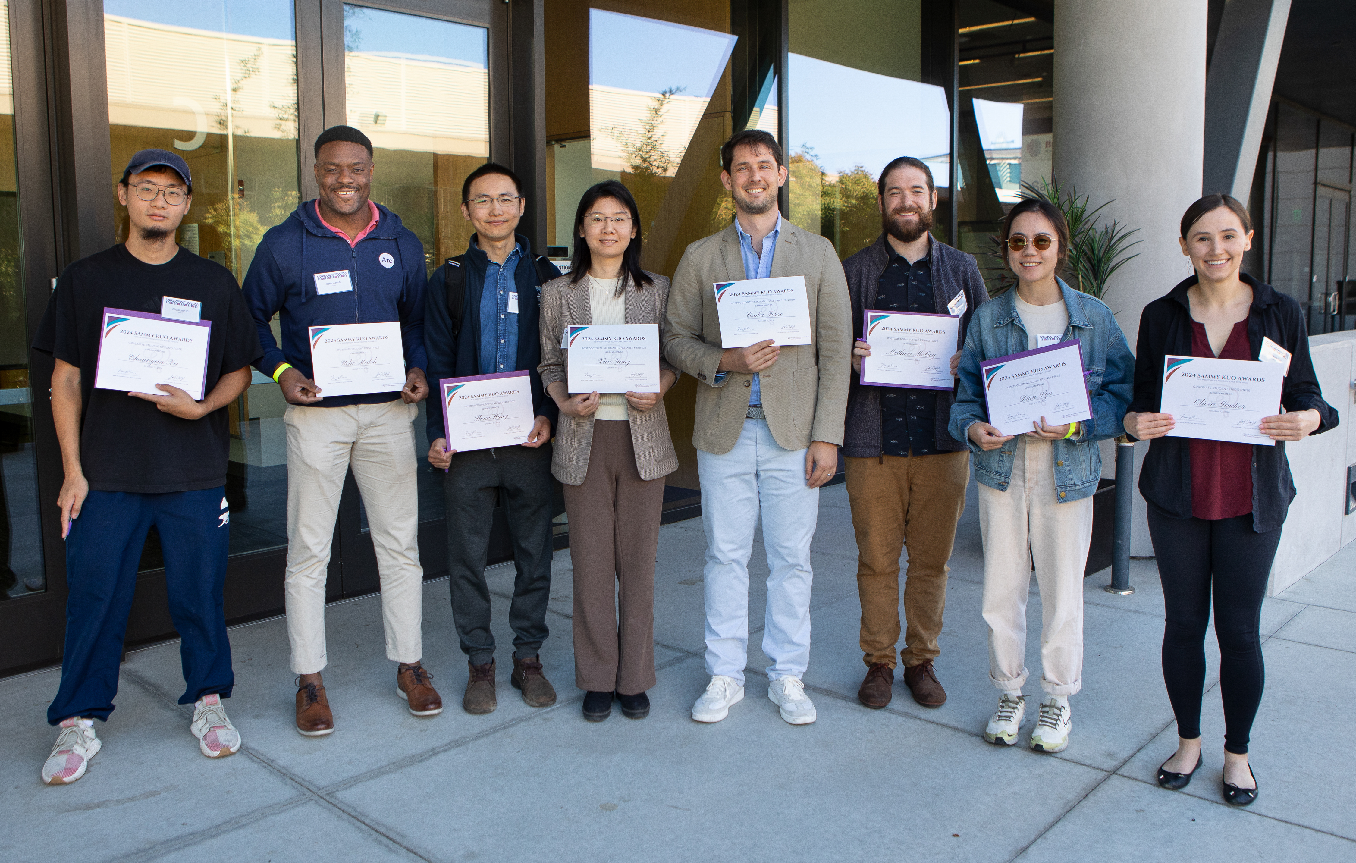 A group of eight individuals standing outside the LKSC building, each holding certificates. From left to right: Chuanyun Xu, Uche Medoh, Shuai Wang, Xiao Yang, Csaba Forro, Matthew McCoy, Chuchu Wang, and Olivia Gautier. All are smiling, and the backdrop includes glass doors and the building's exterior.