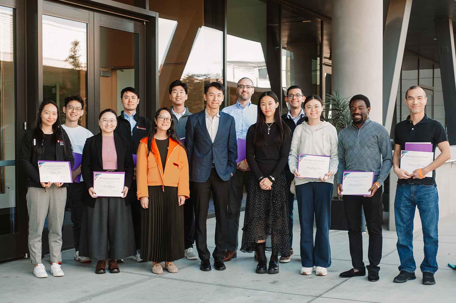 A group of thirteen Sammy Kuo Award recipients and relatives of the late Sammy Kuo stand together outside a building. Kang Shen stands at the center. Several awardees hold certificates. Everyone is smiling and facing the camera. Photo by Ola Hopper.