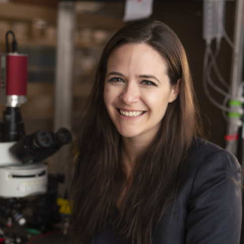 A person with long hair standing beside a microscope in a laboratory setting.
