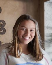 A young woman with long, light brown hair smiles warmly at the camera, wearing a white sweater.