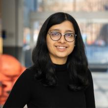 A professional headshot of Aditi Jha, smiling while wearing glasses and a black sweater. She stands in a brightly lit indoor space with blurred windows and a red sculpture in the background.