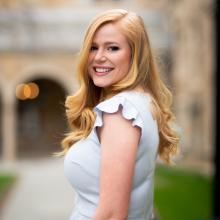 A professional headshot of Annie Goettemoeller, smiling and standing outdoors. She is wearing a light-colored blouse, and the background features blurred architecture and greenery for a natural and elegant feel.