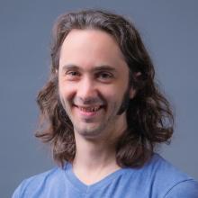 A professional headshot of Jeremy Axelrod, smiling and wearing a blue V-neck shirt. The background is a neutral gray, with Jeremy’s long wavy hair adding a casual and approachable touch.