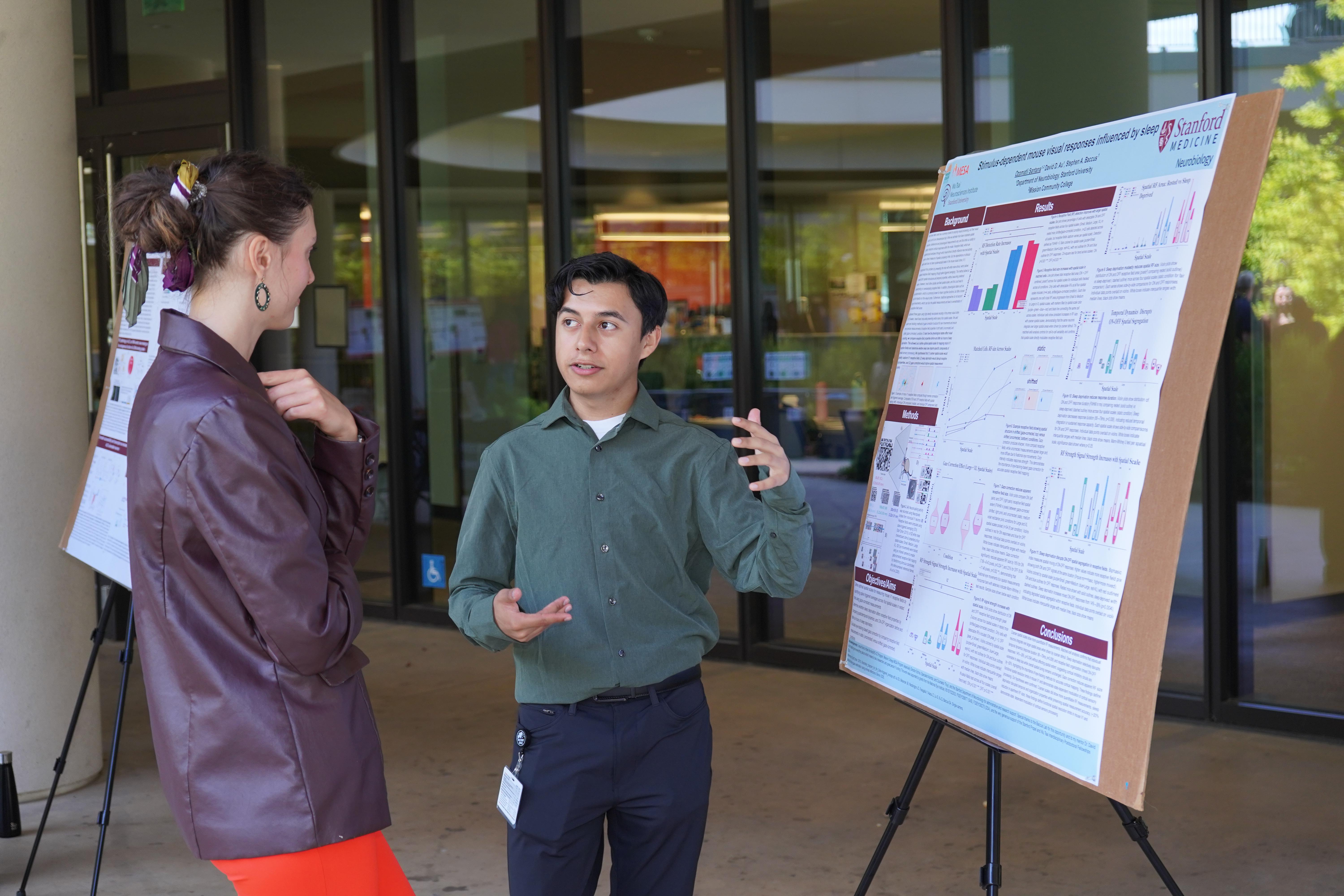 Ozomatli Santana speaks to another person in front of a research poster in a courtyard.