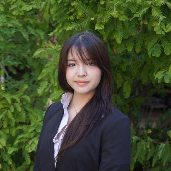 Jillian Quach, wearing a black blazer, stands in front of green foliage and looks to the camera