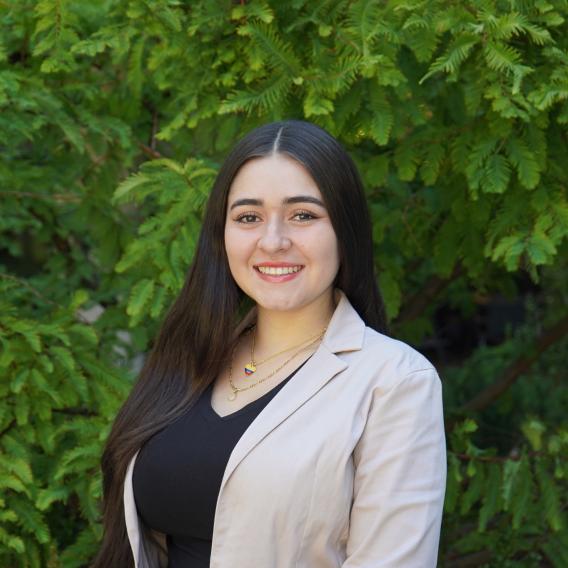 Natalia Jaramillo Becerra, wearing a black top and a tan blazer, stands in front of green foliage and smiles to the camera.