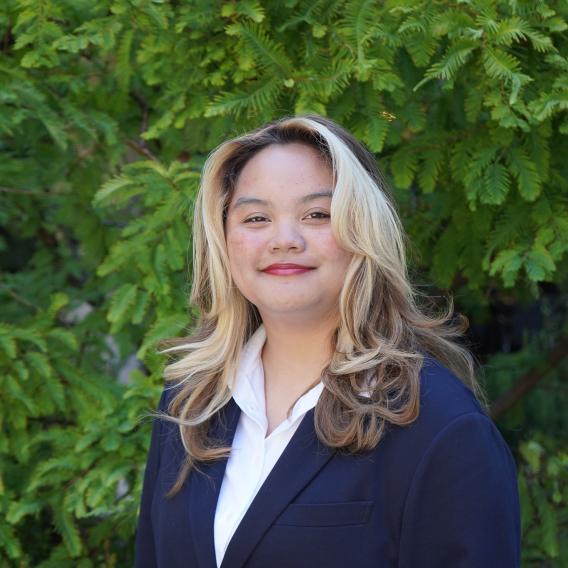 Aljona Reyno, wearing a black blazer, stands in front of green foliage and smiles to the camera.