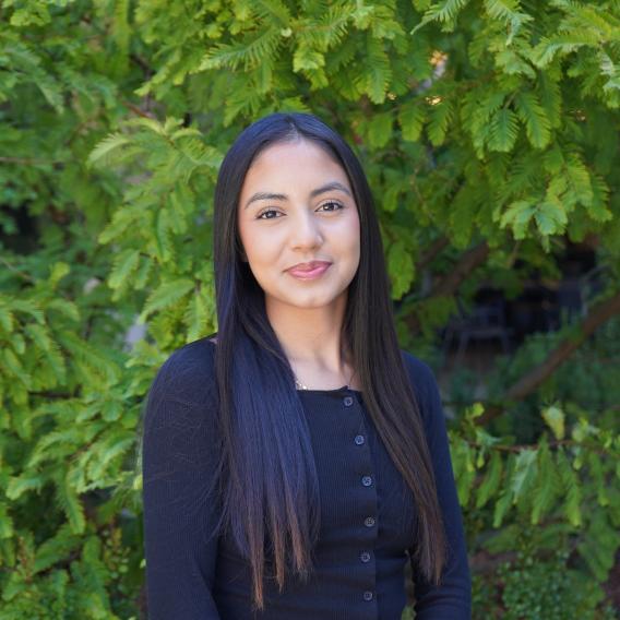 Emely Cisneros wears a black top and stands in front of green foliage while smiling at the camera.