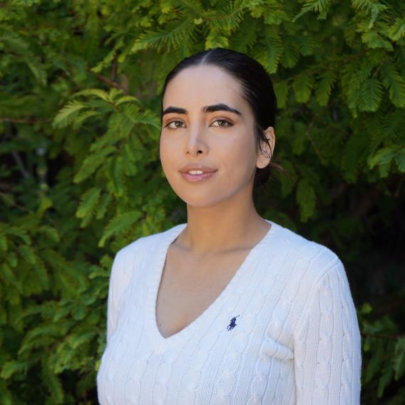 Momina "Muina" Zaman wears a white top while standing in front of green foliage and looks to the camera.