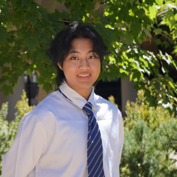 Jaemon Jumpawong wears a button-up shirt and a tie while standing in front of green foliage and smiles at the camera.