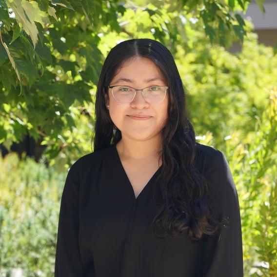 Stefani Garza Barillas wears a black top and stands in front of green foliage while smiling at the camera.