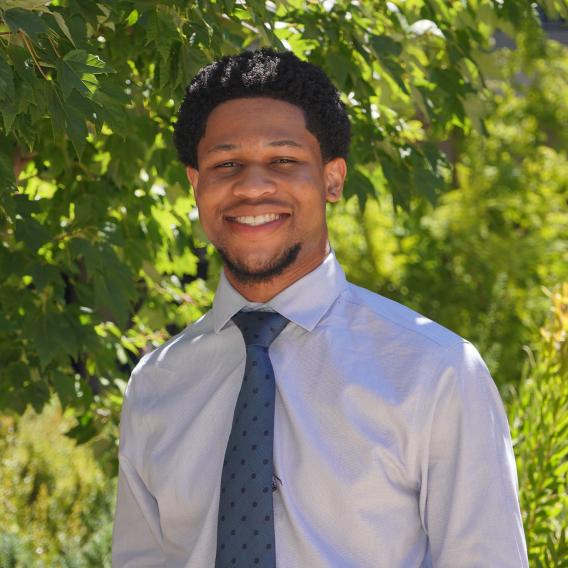 Samuel Egboh stands in front of green foliage, wearing a button-up shirt with a tie, and smiles at the camera.