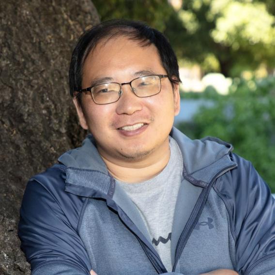 Image of Xiaojie Qiu, who is wearing glasses and a grey-blue hoodie while leaning against a tree and smiling at the camera.