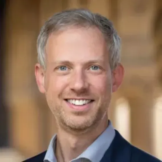 Cory Shain smiles at the camera in front of a blurred backdrop of Stanford architecture