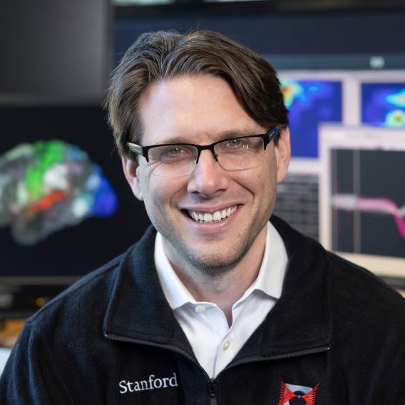 Frank Willett, wearing glasses, a white button-up shirt, and a black jacket, smiles at the camera. In the background are various computer screens showing brain imaging