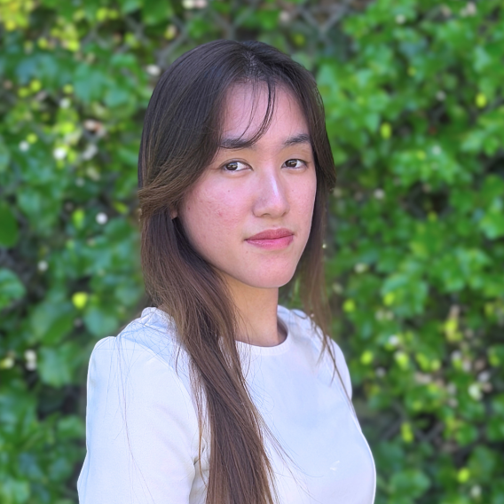Head-and-shoulders portrait of a person with long brown hair wearing a white blouse, posed outdoors in front of a sunlit green hedge.
