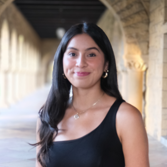 Andrea Roman-Fernandez standing in Stanford sandstone archway wearing a black one-shoulder top and necklace, upper body visible.