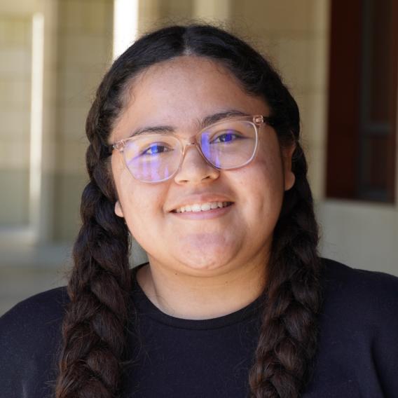 Head-and-shoulders portrait of Denise with two long braided pigtails wearing a black crewneck, photographed in a Stanford sandstone archway; face is obscured.