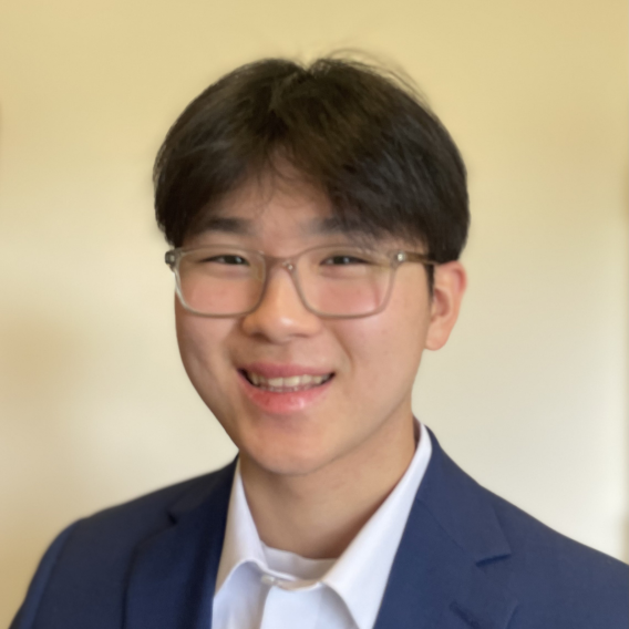 Head-and-shoulders portrait of Hayden Kang wearing a navy blazer, white collared shirt, and glasses against a plain beige background.