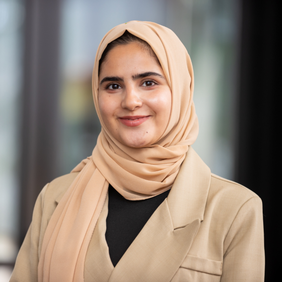 Husna Ayoubi wearing a beige hijab and matching blazer over a black top, posed against a softly blurred indoor background.