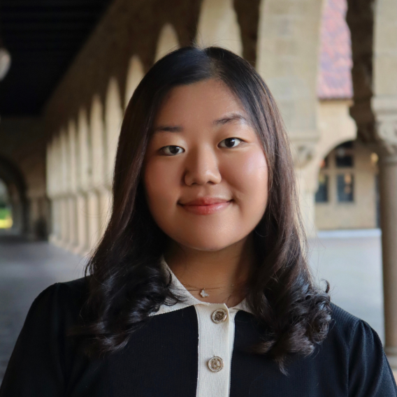Jamie Kwon wearing a black sweater with a white buttoned collar and necklace, standing in Stanford sandstone archway with columns visible behind them.
