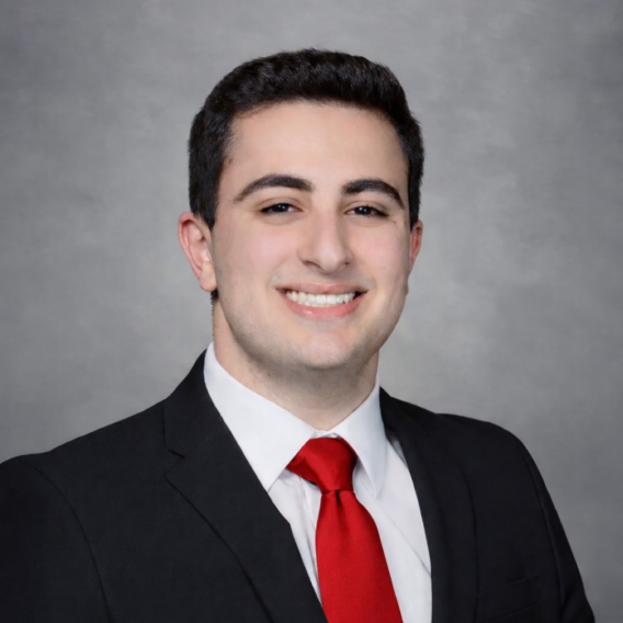 Head-and-shoulders portrait of Joshua Barsoian wearing a dark suit, white shirt, and red tie against a neutral gray studio background.