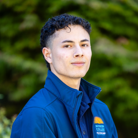 Head-and-shoulders portrait of a person with short curly hair wearing a blue zip-up jacket featuring a Mission College "Welcome Center" logo, posed outdoors against a blurred green background.