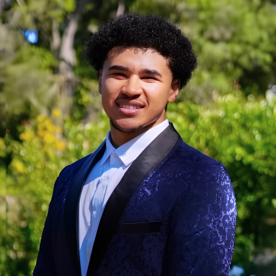 Head-and-shoulders portrait of a person with a short curly hair wearing a dark blue crushed-velvet tuxedo jacket with satin lapel and a white dress shirt, posed outdoors against green foliage.