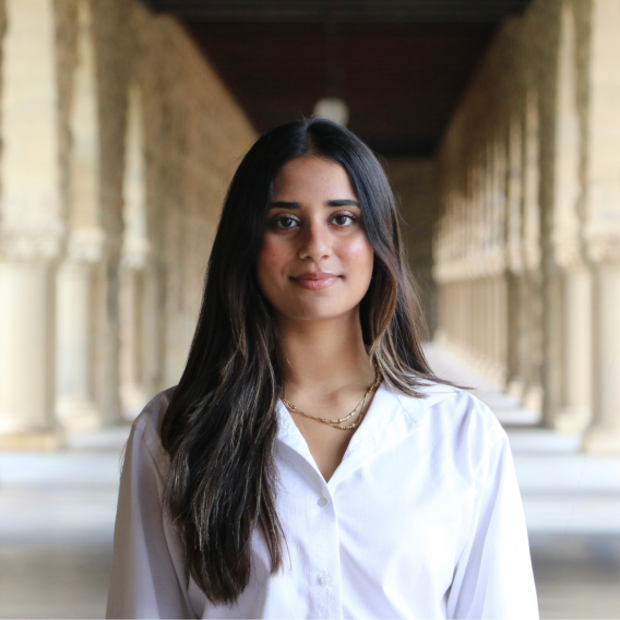 Malaika Naseer standing under Stanford sandstone archway wearing a white button-down shirt and layered gold necklaces, long dark hair falling past the shoulders.