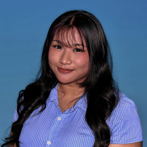 Head-and-shoulders portrait of a person with long wavy dark hair wearing a blue-and-white striped button-up shirt and thin necklace against a solid blue background.