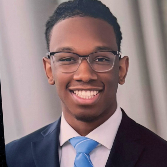 Head-and-shoulders portrait of Milton Jennings wearing a dark suit, white shirt, and light blue tie against a neutral curtain backdrop.