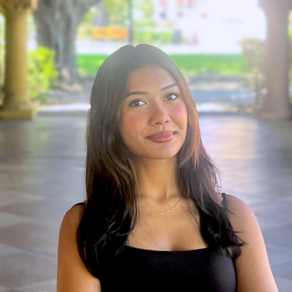 Najifa Nosin in Stanford sandstone archway wearing a black tank top and thin gold necklace, long dark hair falling past the shoulders, upper body visible.