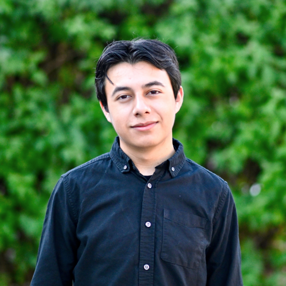 Head-and-shoulders portrait of a person wearing a dark button-down shirt standing outdoors in front of a green leafy hedge.