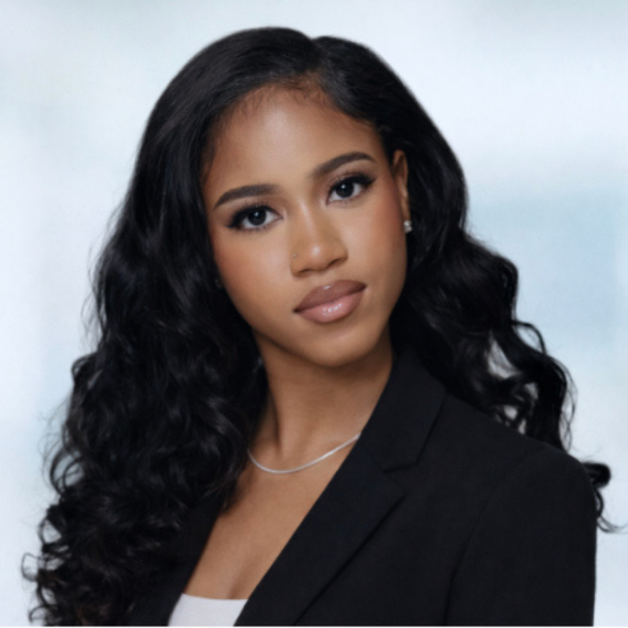 Head-and-shoulders portrait of Talia Dixon with long curly dark hair wearing a black blazer, white top, and thin silver necklace against a soft blue-gray background.