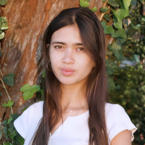 Head-and-shoulders portrait of a person with long brown hair wearing a white short-sleeve top and delicate necklace, posed against a tree trunk with ivy and leafy background.