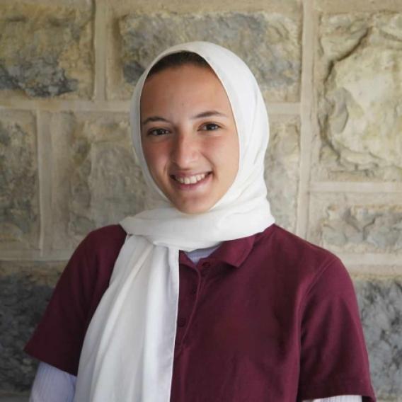 Headshot of Nour Omar wearing a white headscarf and burgundy top, standing in front of a brick backdrop.