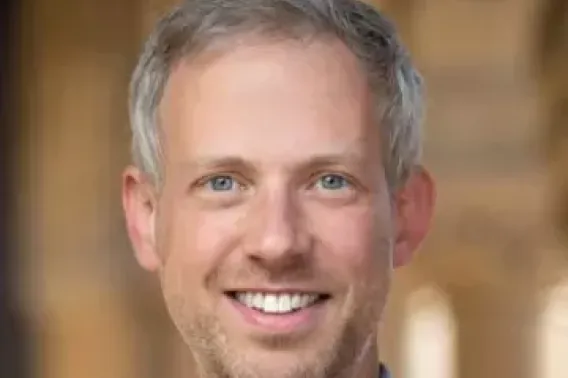 Cory Shain smiles at the camera in front of a blurred backdrop of Stanford architecture