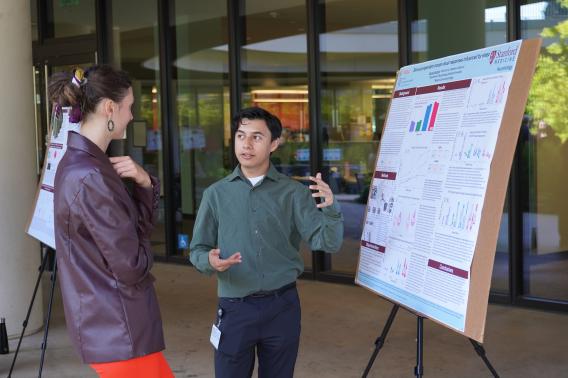 Ozomatli Santana speaks to another person in front of a research poster in a courtyard.